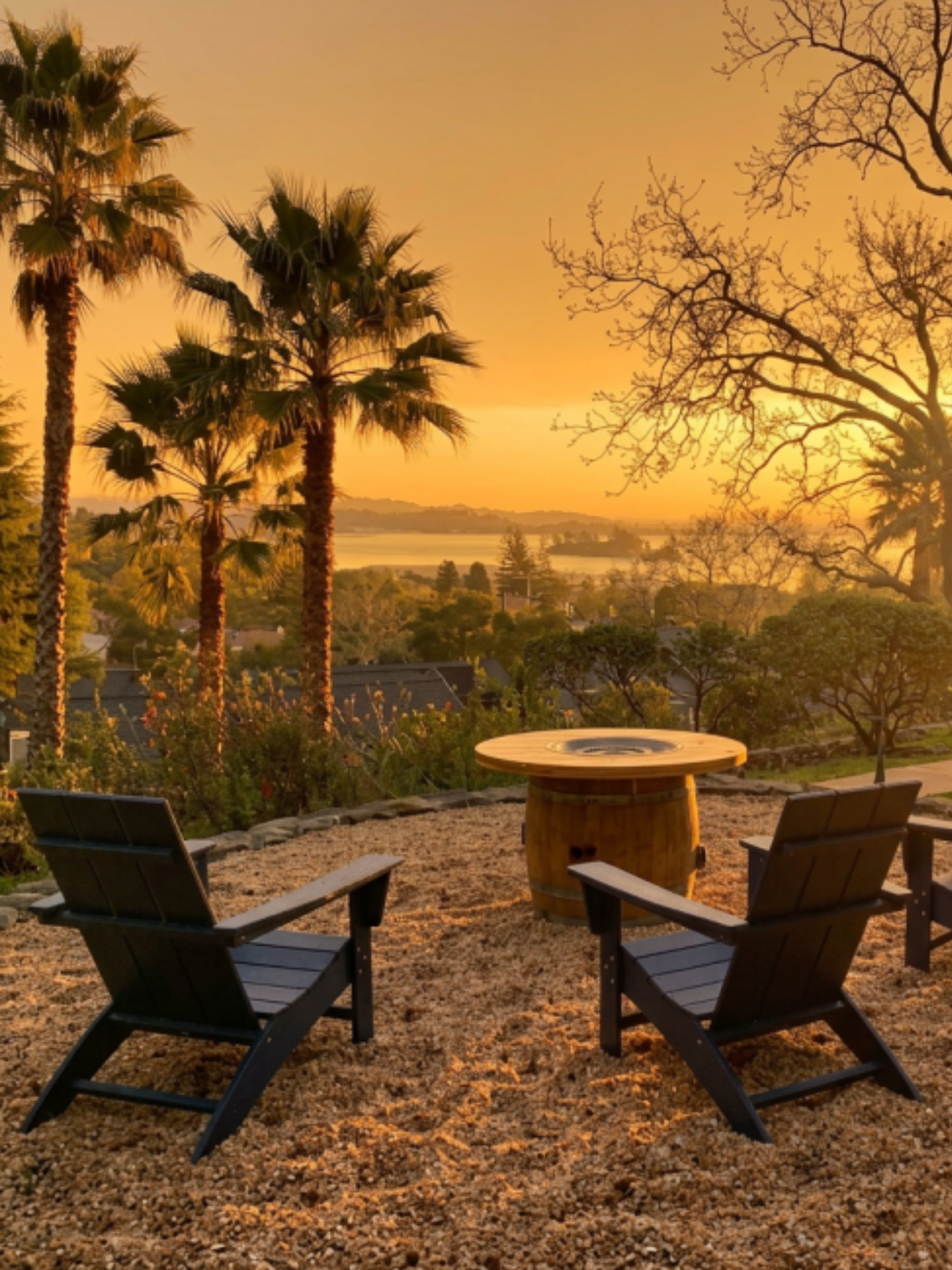 Outdoor seating area with four black chairs around a barrel-style fire pit, overlooking a sunset landscape with palm trees and a body of water in the distance.