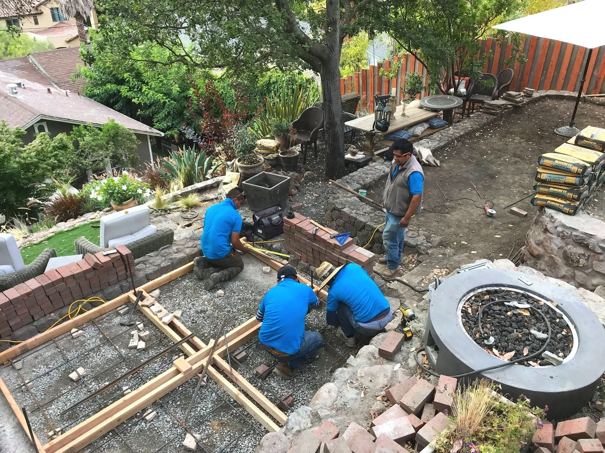 Workers building a brick wall on an outdoor patio area surrounded by plants, trees, and outdoor furniture. Construction materials and tools are visible.