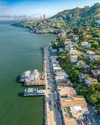Aerial view of a coastal city with a long pier extending into the water, with houses and buildings along the shoreline and hills in the background.