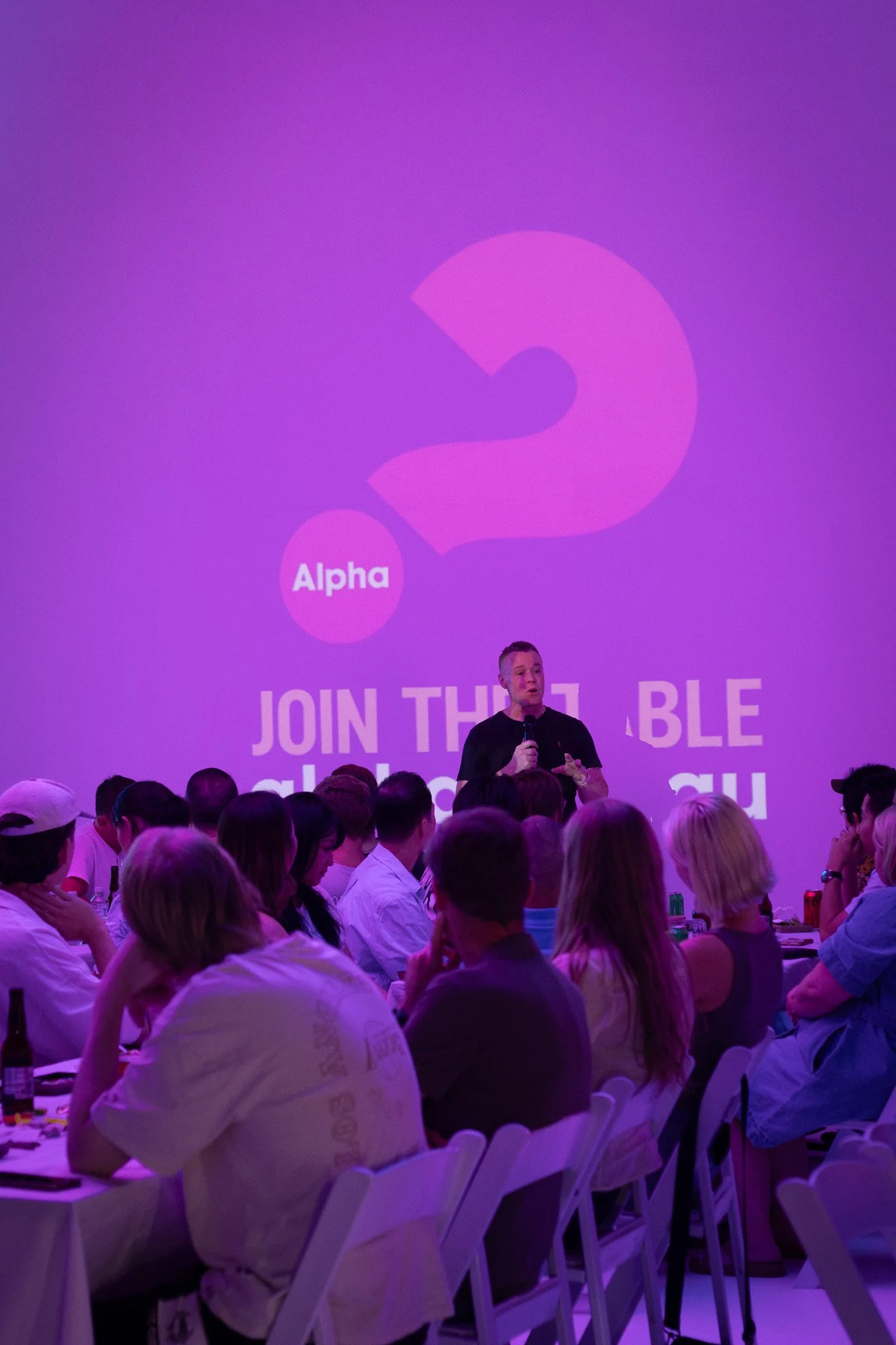 A speaker standing in front of a large purple screen with the text 'Alpha' and a question mark, addressing an audience at a conference or event.