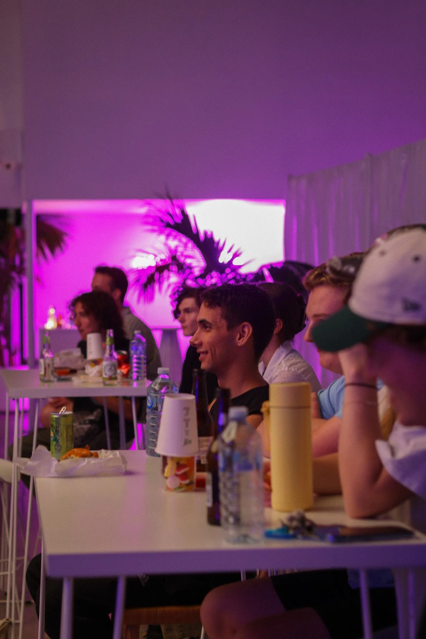 Group of young people sitting at a long white table at a party or event, with beverages and snacks, purple lighting, and a screen displaying palm trees in the background.