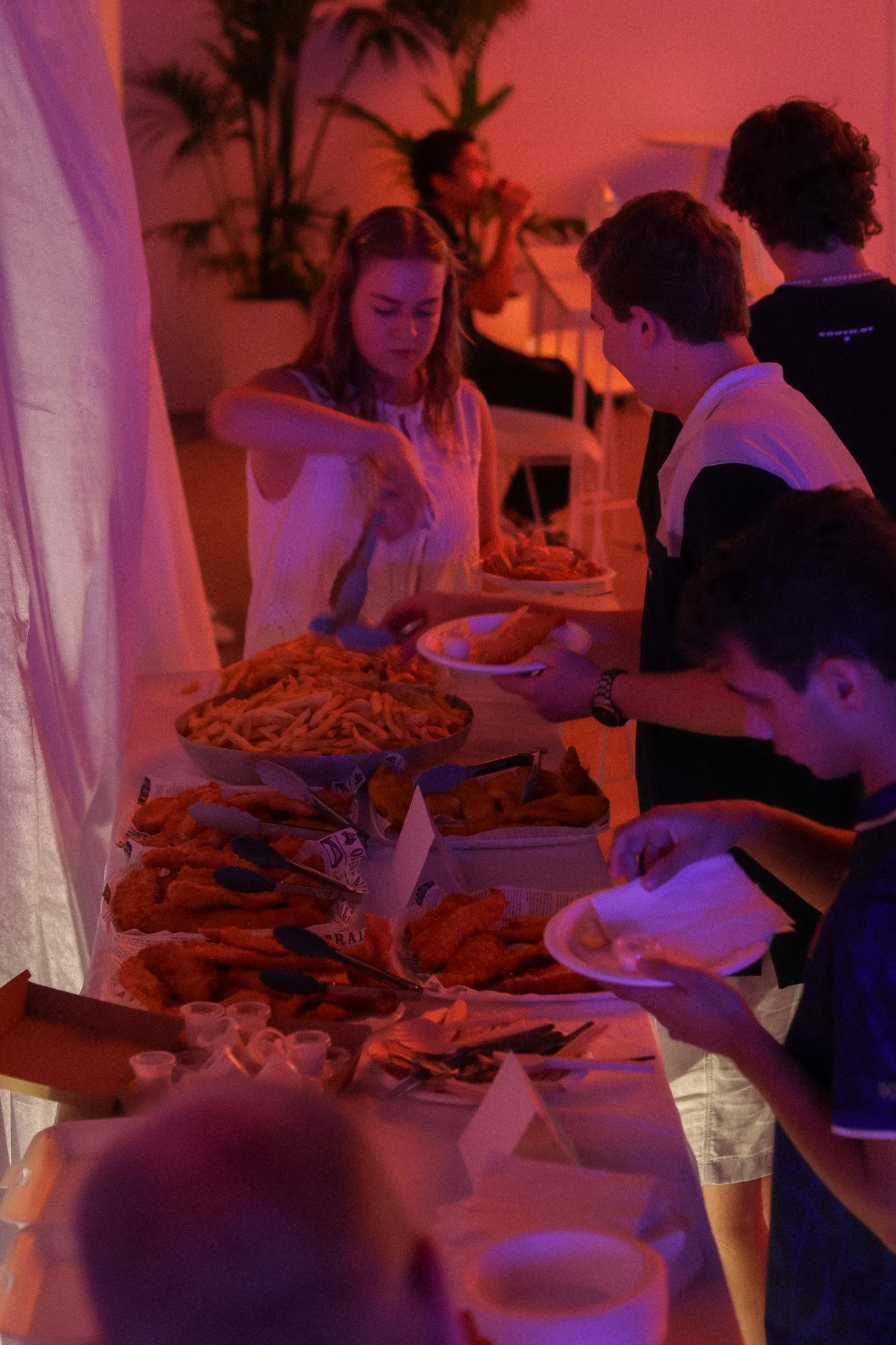 People serving and selecting food at a buffet table with pasta, chicken, and various sides, under pink and purple lighting.