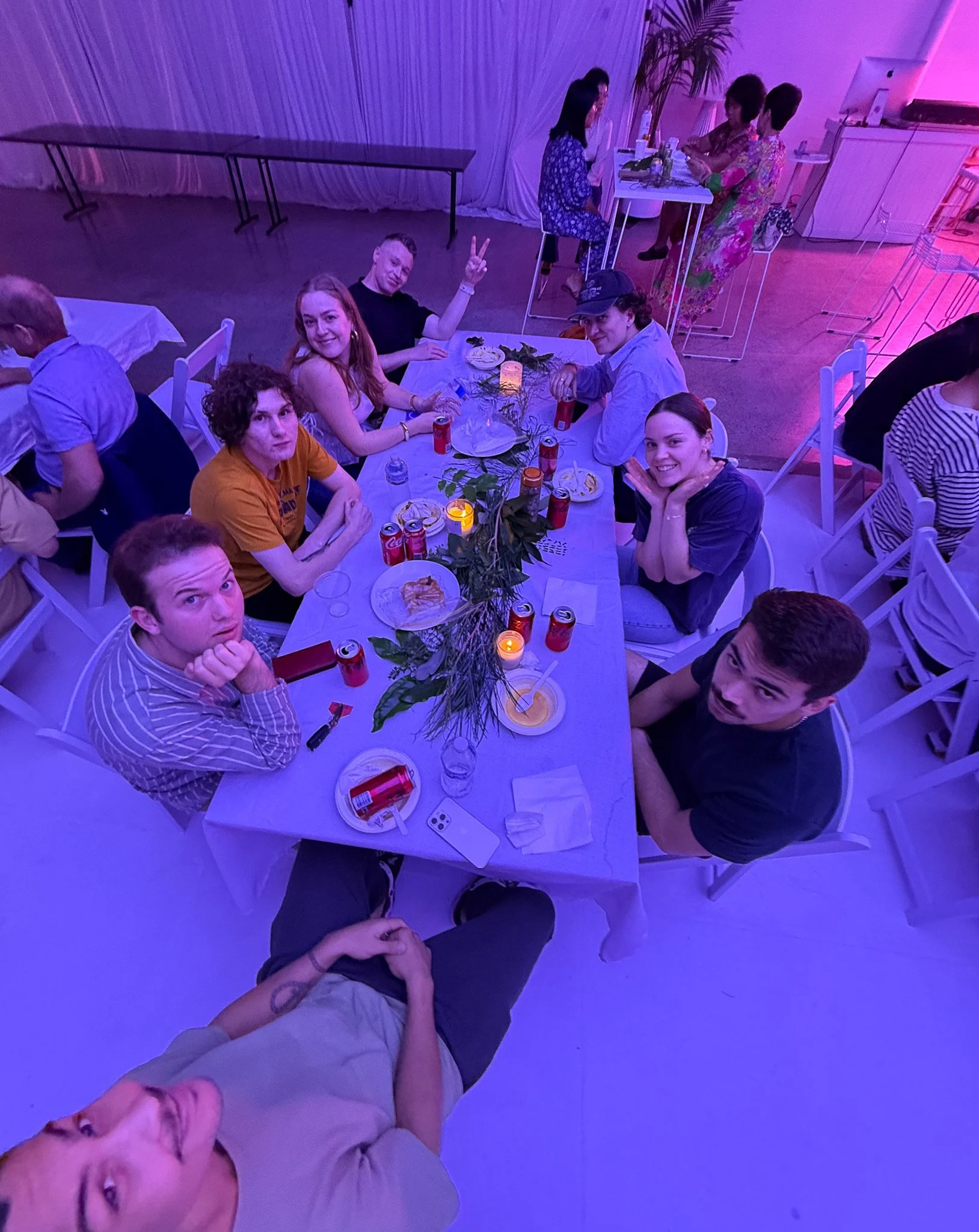 Group of friends sitting around a table at a party, with some making selfies and others eating, in a room with purple lighting and a decorated table.