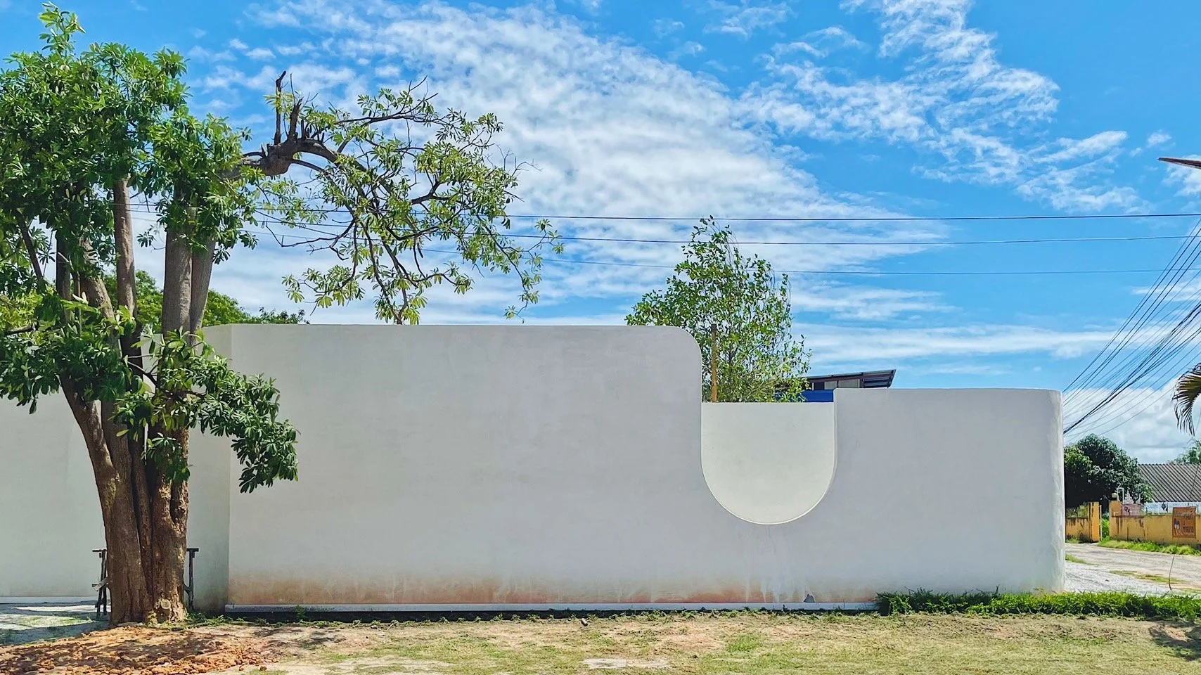 A white wall with a rounded cutout, outdoors under a partly cloudy blue sky. Trees and houses are visible in the background.