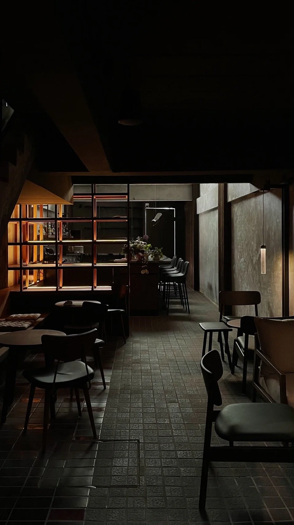 Interior of a dimly lit, modern restaurant or cafe with chairs, tables, and a bar area with stools, featuring a contemporary design with warm wooden accents and textured walls.