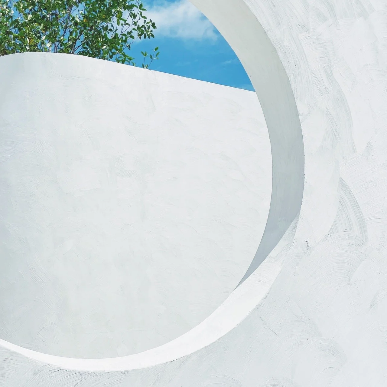 A white architectural structure with curved walls and a circular opening, with a tree and blue sky visible through the opening.