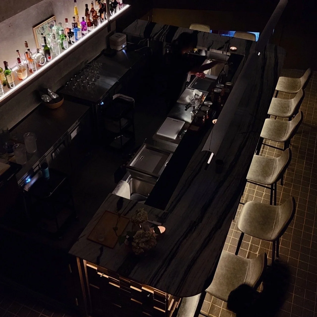 A dimly lit bar with a long dark wooden counter. Several beige chairs are lined along one side for patrons. Behind the bar, a shelf holds various bottles of alcohol illuminated by backlighting. A person is working behind the counter, and there are some bar tools and glassware on the counter.