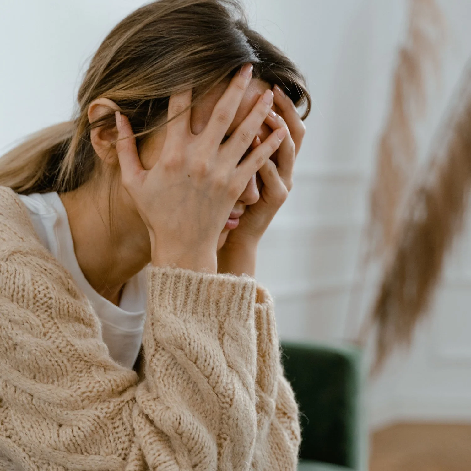A woman with brown hair wearing a beige knit sweater, holding her head with both hands and appearing distressed or overwhelmed.