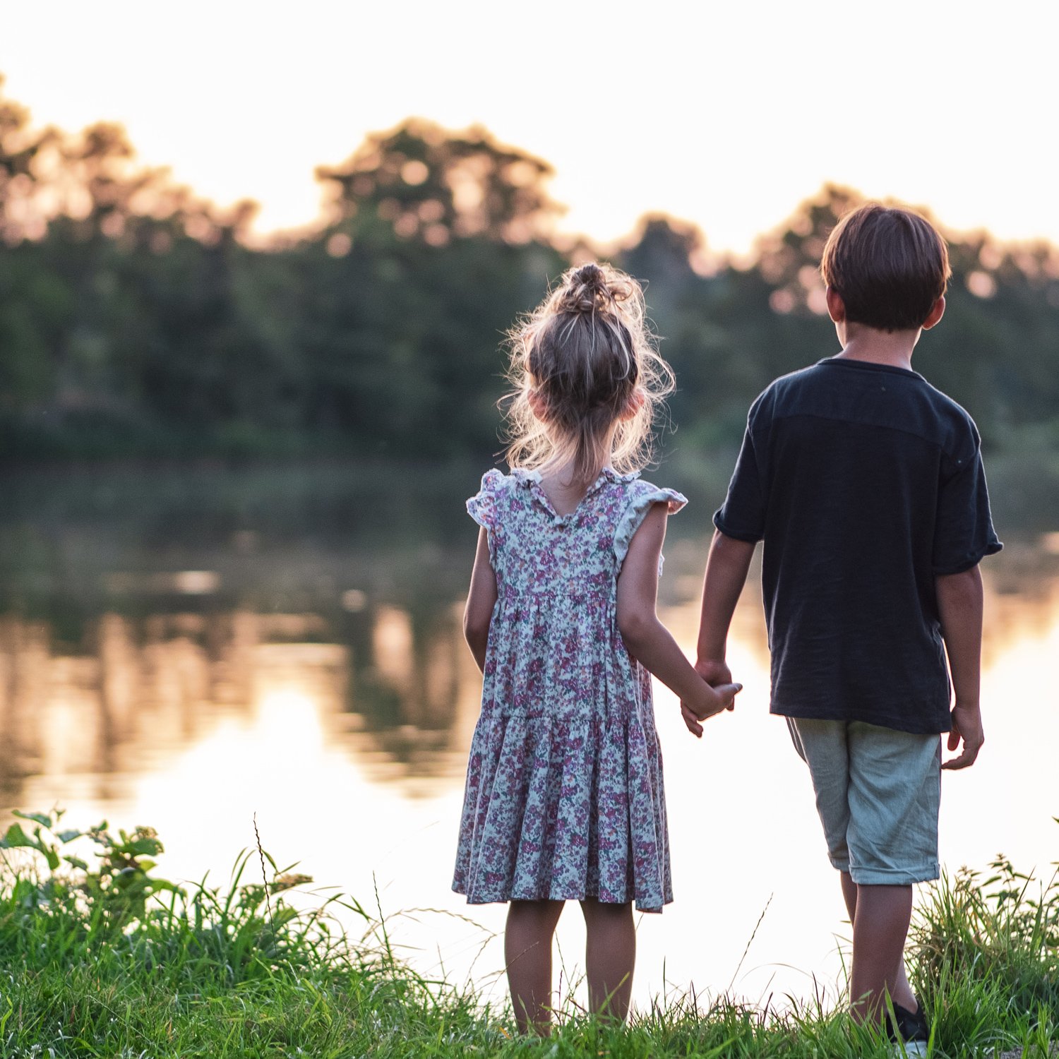 Two children, a girl and a boy, holding hands and standing by a body of water at sunset or sunrise, with trees in the background.