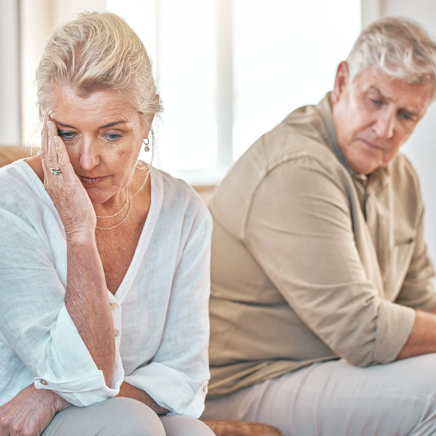 An elderly woman and man sitting side by side, looking down with concerned expressions. The woman is touching her forehead with her hand, and the man is slightly turned away, both appearing worried or contemplative.