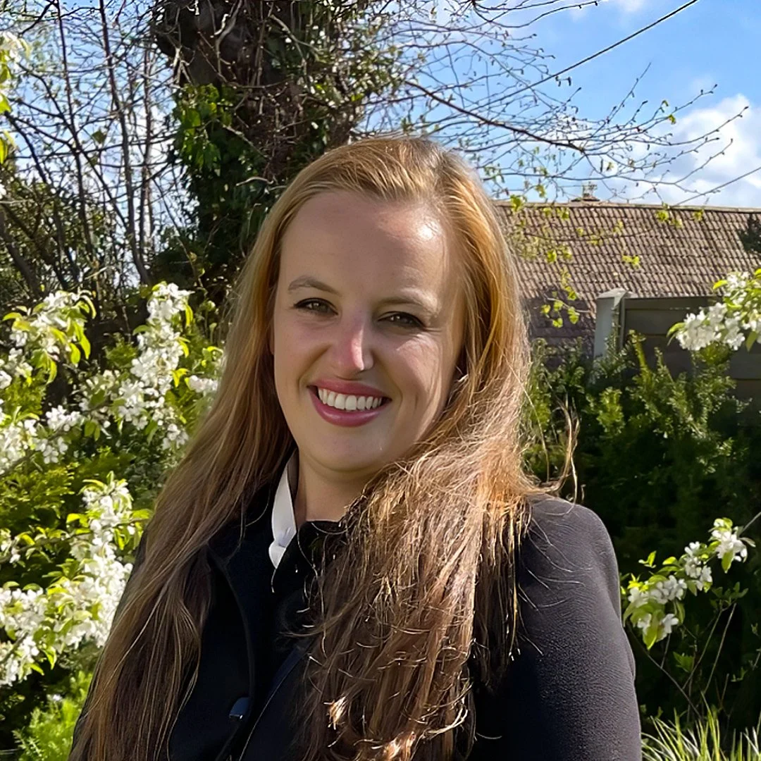 A smiling woman with long, light brown hair standing outdoors in a garden with white flowers and green foliage, under a partly cloudy sky.