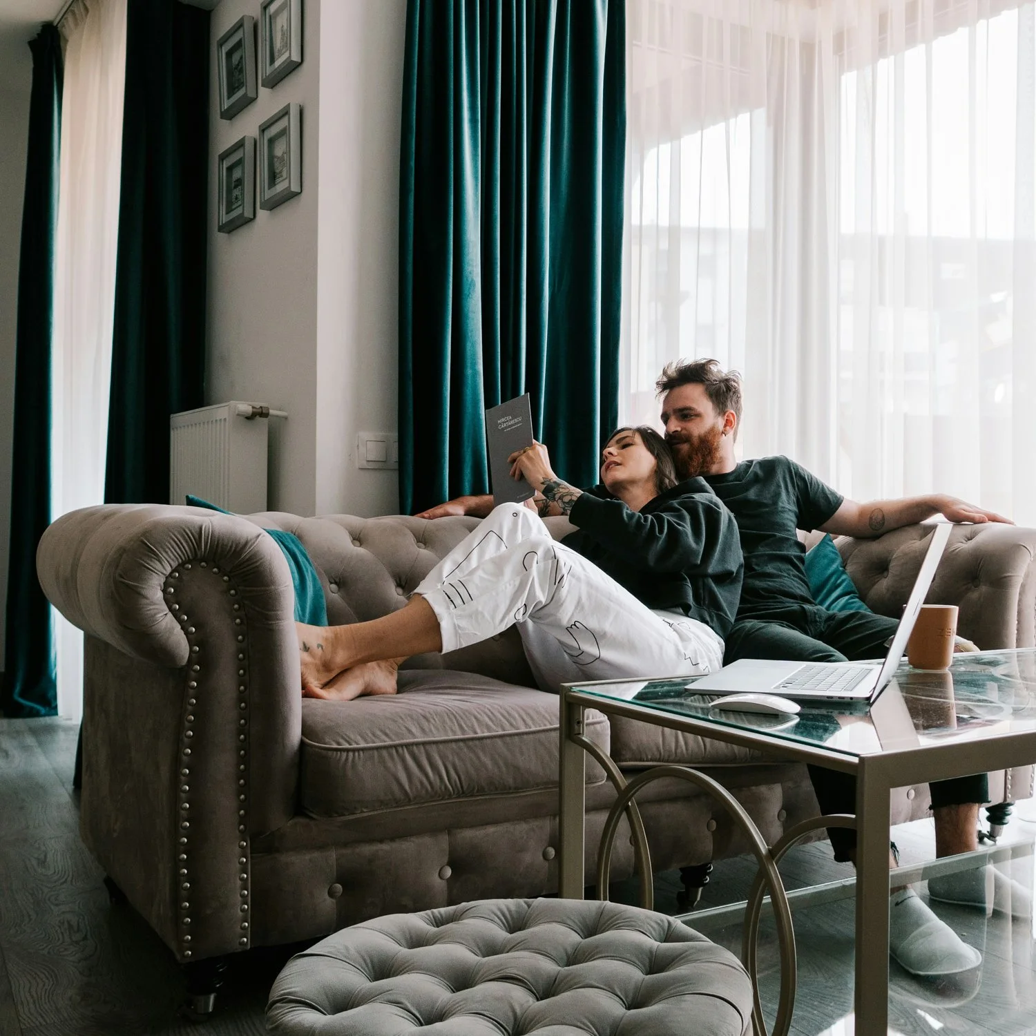 A man and woman relaxing on a beige sofa in a bright living room, with the woman reading a book and both looking comfortable and cozy.