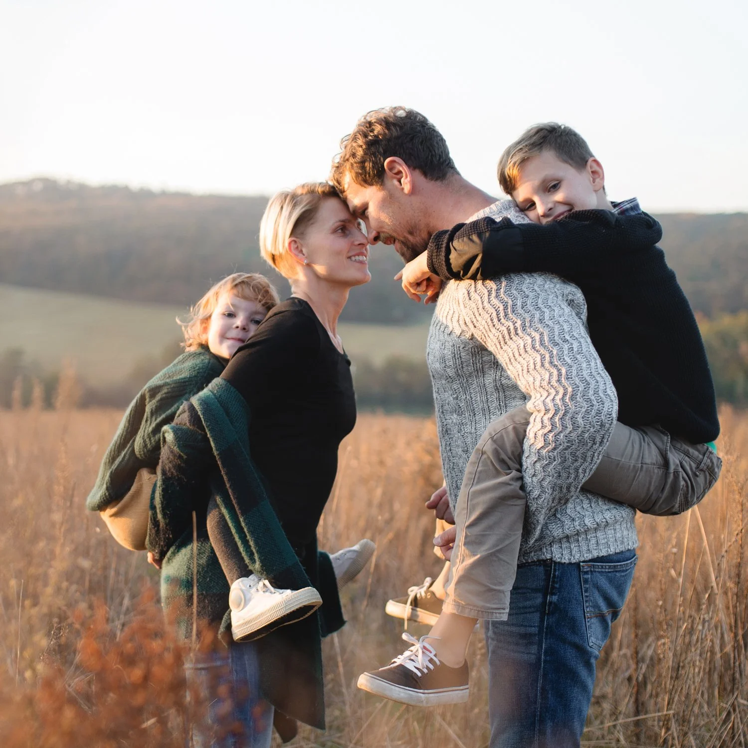 A family of four standing in a field of tall grass outdoors, with mountains in the background, smiling and touching foreheads, during what appears to be late afternoon or early evening.