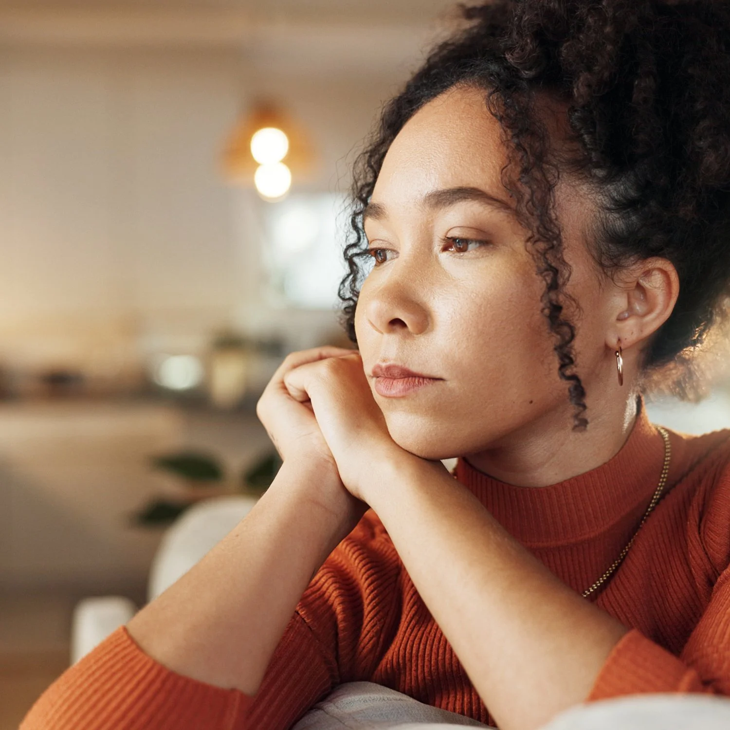 A young woman with curly dark hair, wearing a burnt orange turtleneck sweater, resting her chin on her clasped hands and gazing thoughtfully to the side in a warmly lit indoor space.