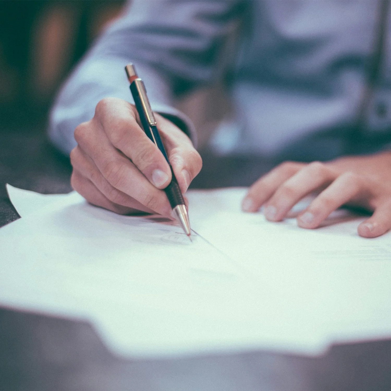 A person wearing a white shirt is writing on a sheet of white paper with a black and silver ballpoint pen.