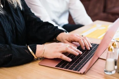 Two people working on a rose gold laptop at a wooden table, with one person's hands typing and the other's seated nearby.
