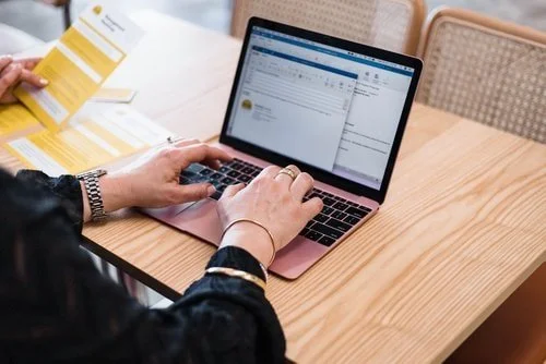 Person typing on a laptop at a wooden table, with printed documents nearby.