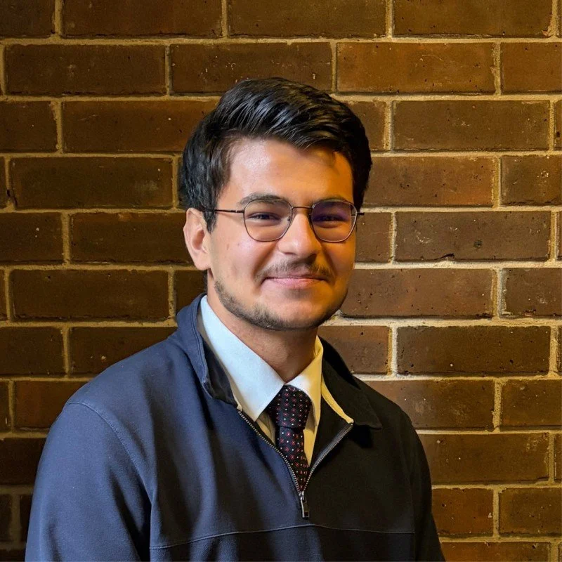 Young man in suit, standing in front of a blue background with white emblem featuring a government building and laurel wreaths.