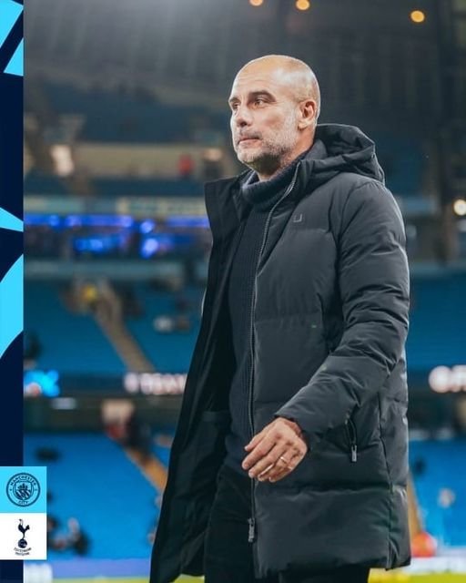 En mann med kort grått skjegg og bald hode, iført svart jakke, står på en fotballbane utendørs.