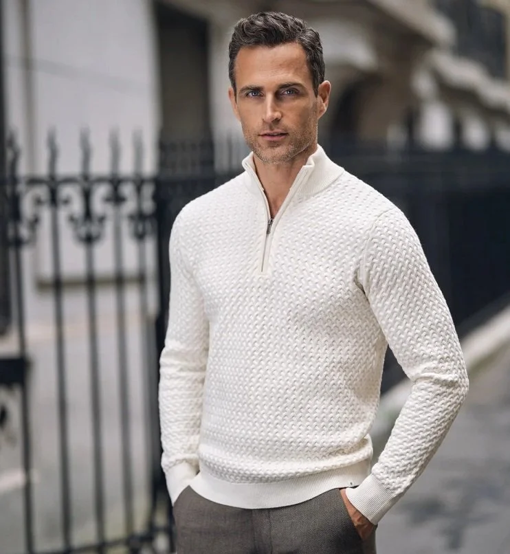 En mann med mørkt, krøllete hår og lys hud står utendørs foran en svart port og gamle bygninger, iført en hvit, teksturert genser med glidelås i halsen og mørke bukser.