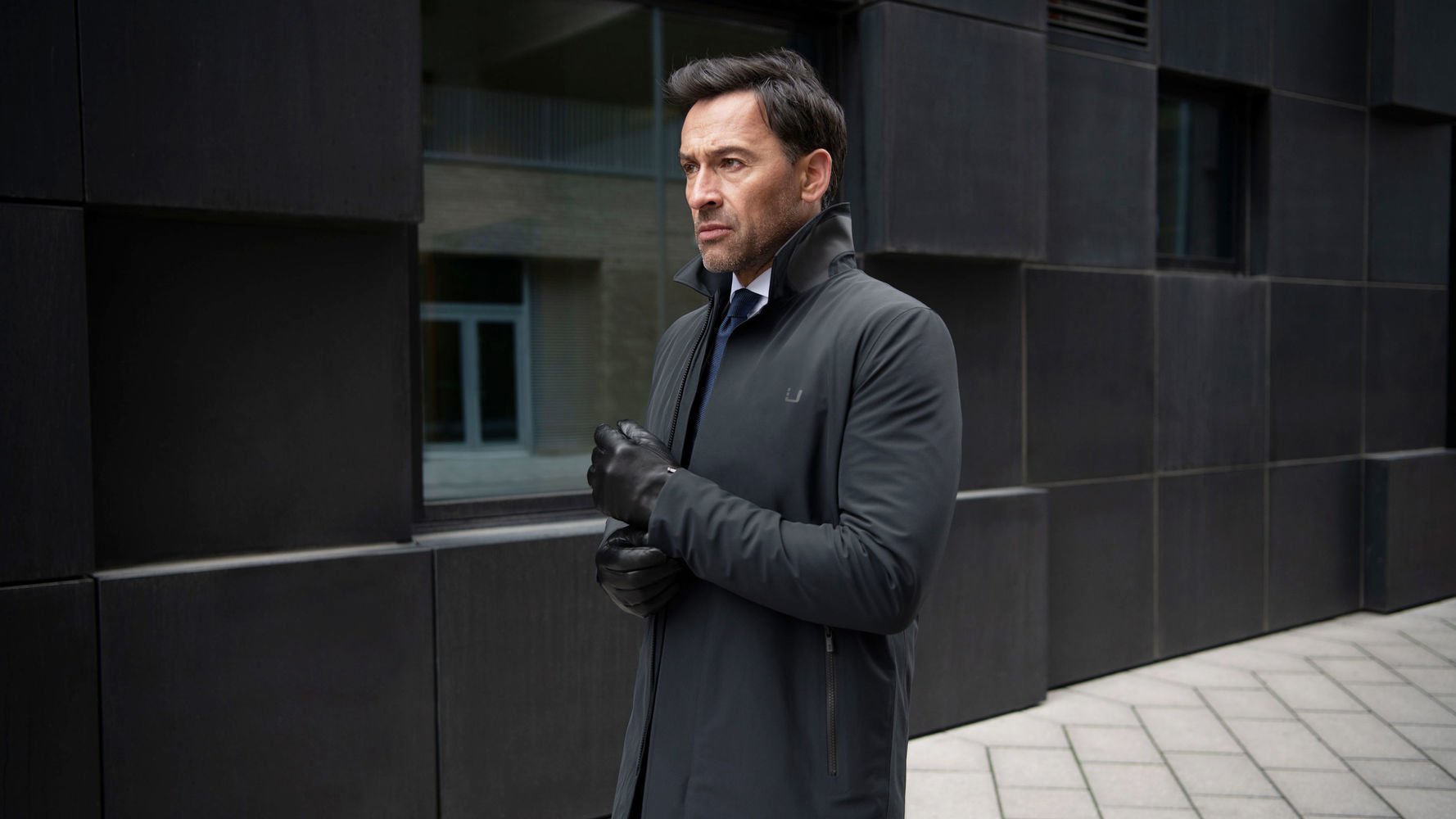 En mann i svart jakke og hansker står utendørs foran en mørk bygning, med en alvorlig uttrykk.