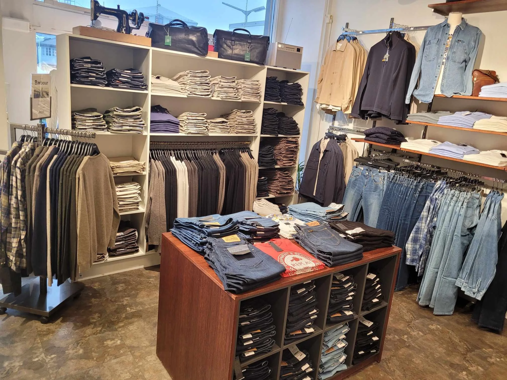 Innvendig bilde av klesbutikk med hyller og plagg av jeans, bukser, skjorter og gensere. Det er også et bord med brettede bukser og jeans i sentrum.