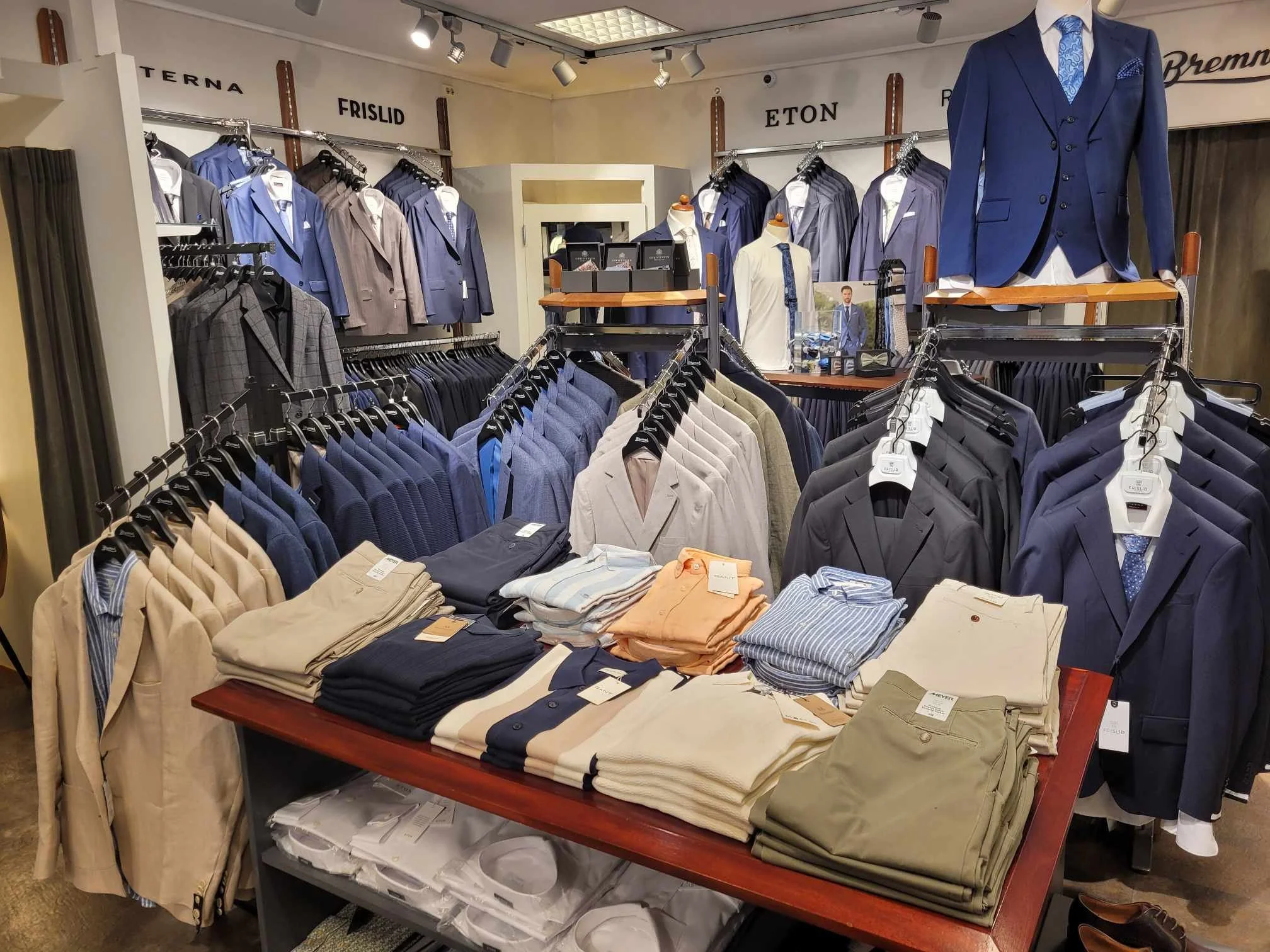 Mannsdrakter og skjorter i et herretøysbutikk. Flere eskester av dresser og skjorter er utstilt på en bord og stativer.