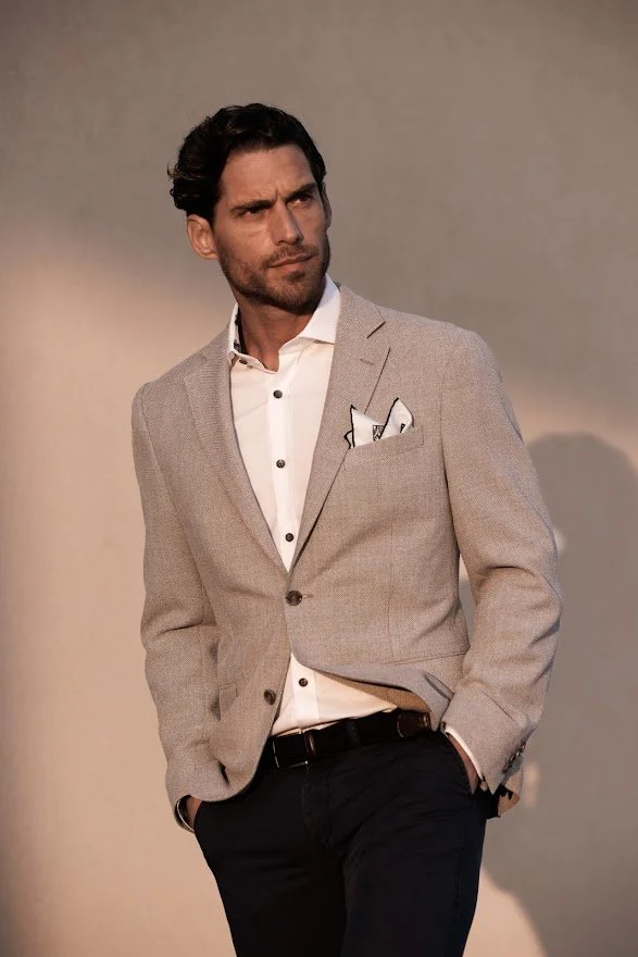 En mann med mørkt, krøllete hår og skjegg, iført en lys grå dressjakke og hvit skjorte, står med hånden i lommen mot en ensfarget bakgrunn.