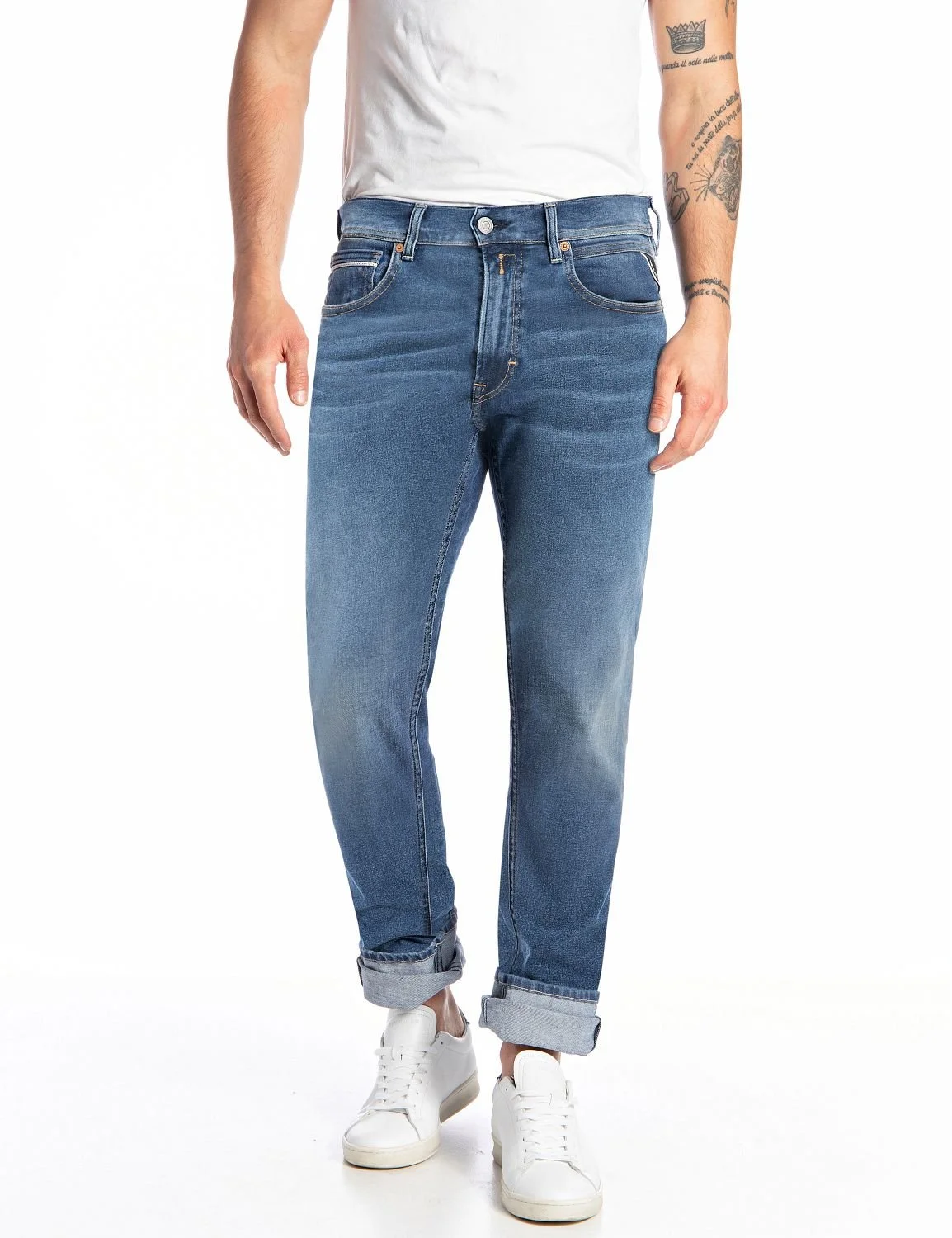 Person iført blå jeans, hvit t-skjorte og hvite sneakers, med synlige armtatoveringer.