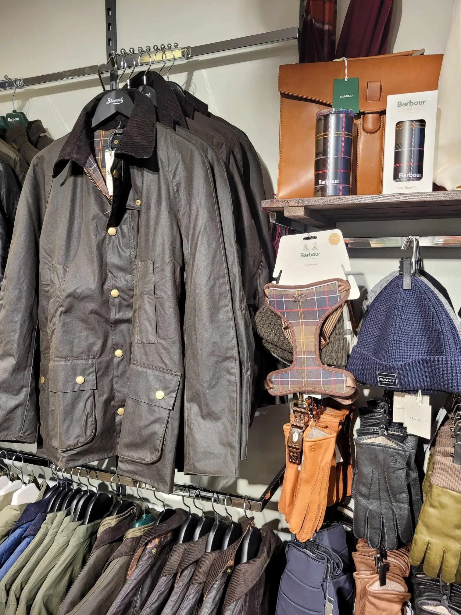 Rekke med jakker, hansker, og tilbehør i en butik, inkludert en grå jakkesak, en blå lue, votter, og termoser.