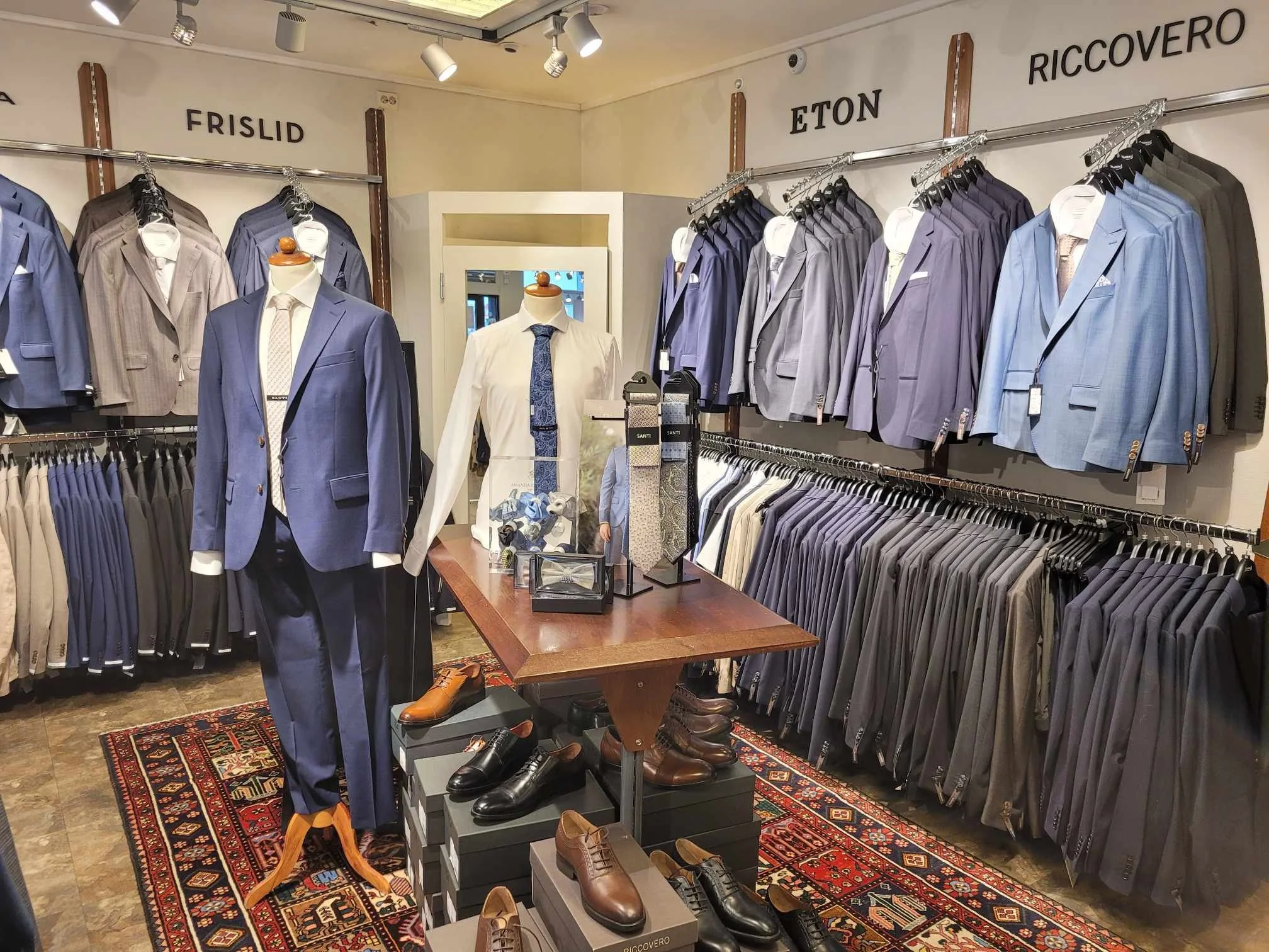 Herrefrakker og kostymer på kles­vise etter butikk. Mannequins iført dresser og sko i fokus. Rader med dresser og jakker på hyller og stativ. Merker som 'FRISLID', 'ETON' og 'RICCOVERO' på veggene.