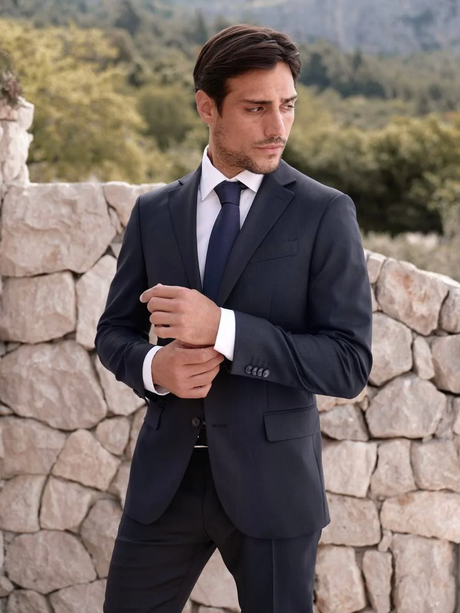 En mann i dress står foran en steinmur med en naturlig landskap i bakgrunnen.