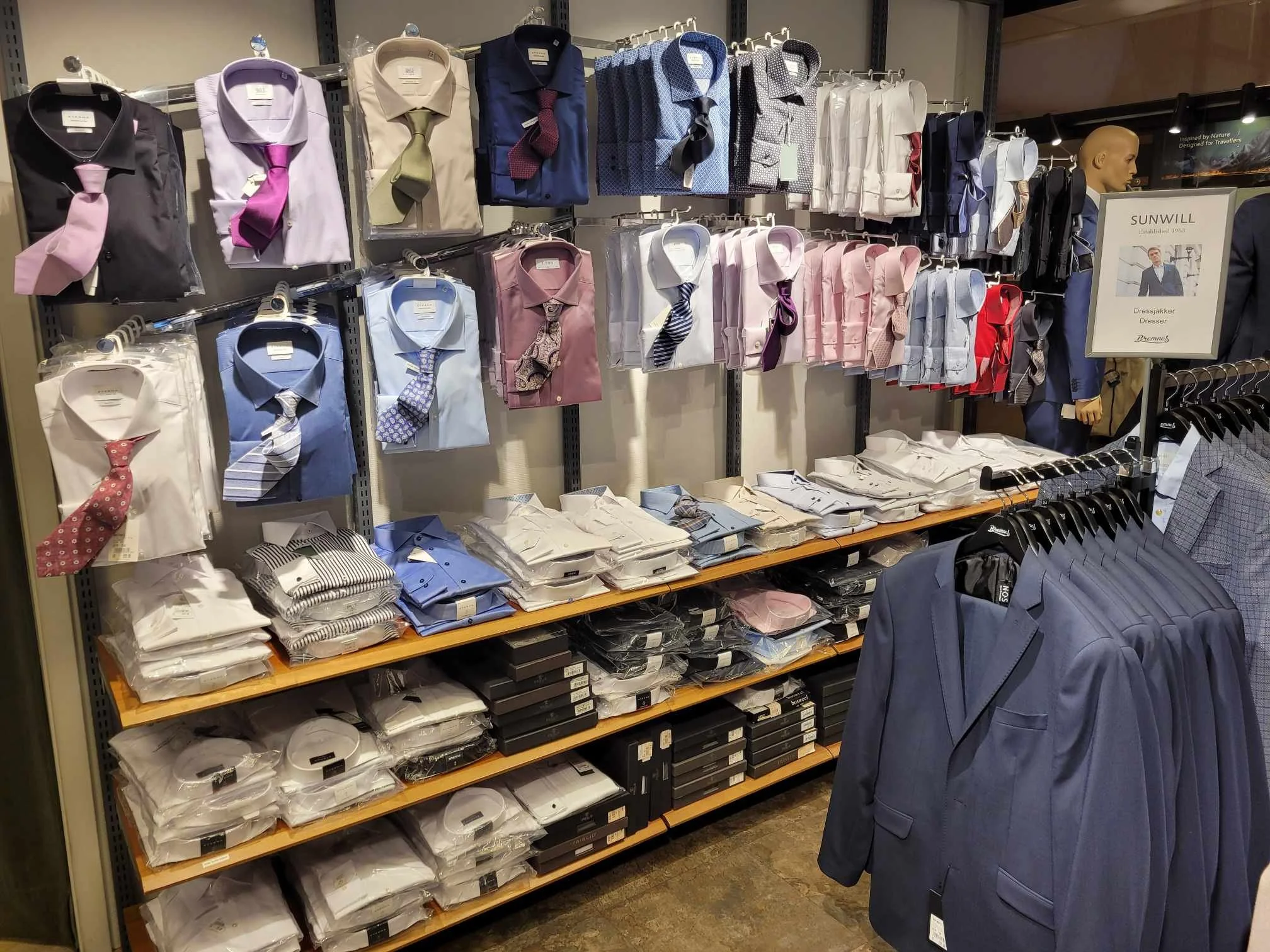 Hylleseksjon med ferdigknyttede slips, skjorter i ulike farger og mønstre, og dressjakker i marineblå og grått, klar til kjøp.