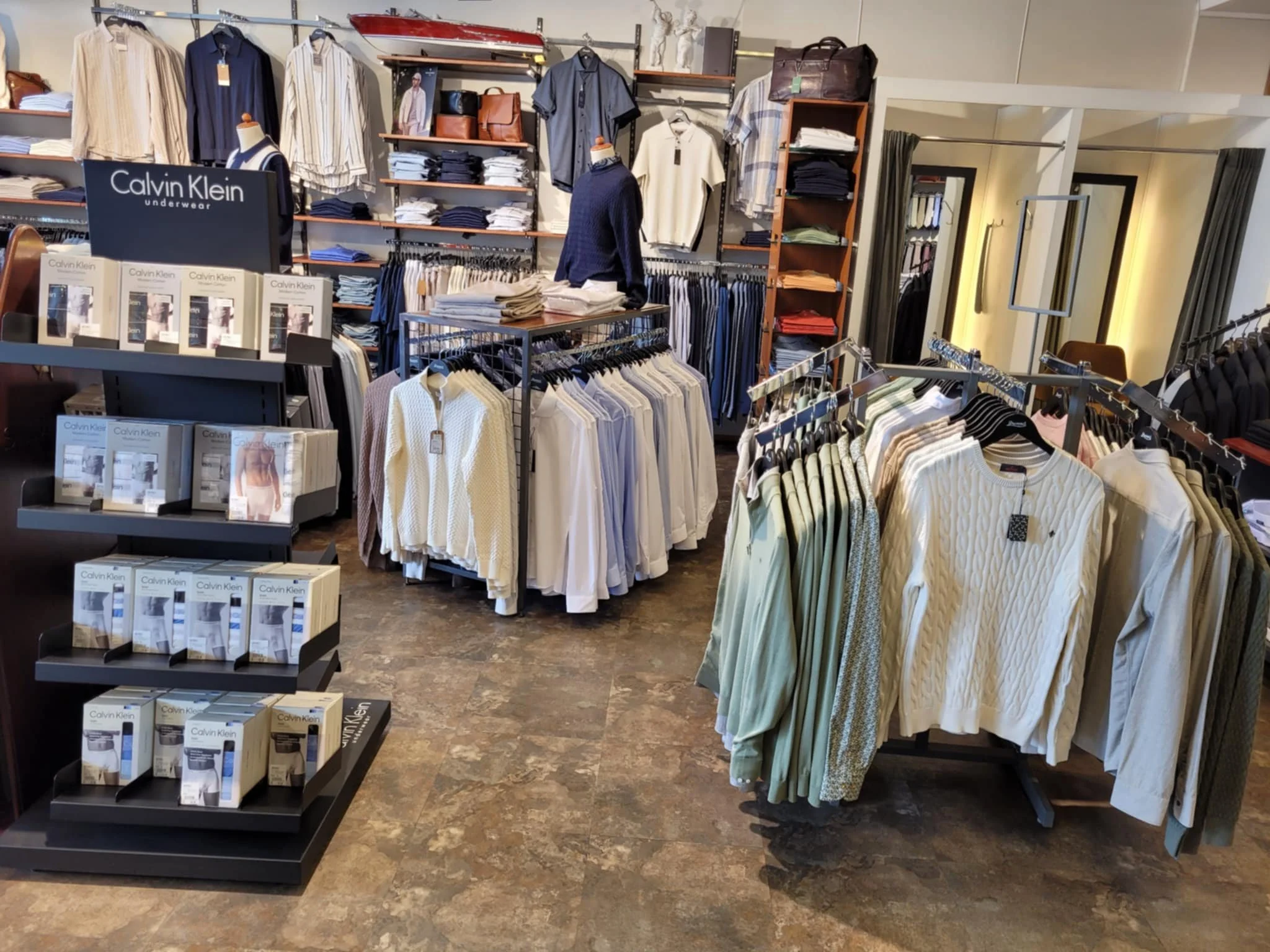 Innendørsbildet viser en klesbutikk med flere grupper av lodne gensere, skjorter og bukser på klærstativ og hyller. Det er plakater for Calvin Klein under undertøy, og det er mannekenger med trøyer og klær i bakgrunnen.