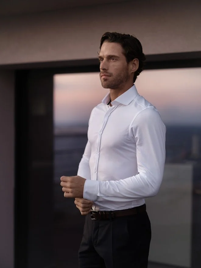 En mann i hvit skjorte står på en balkong med utsikt over en by eller landskap ved solnedgang.