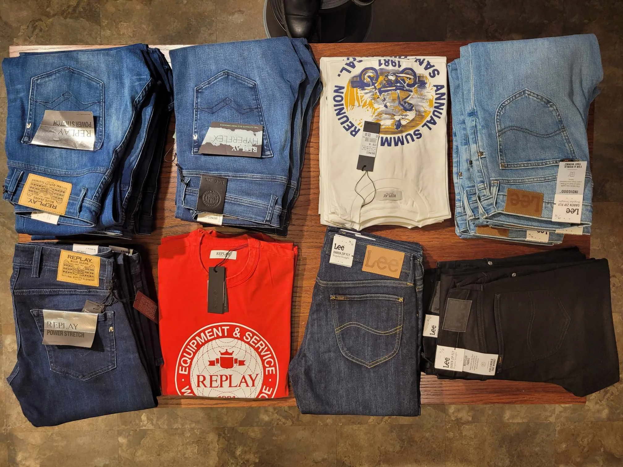 Ulike bukser og en t-skjorte som ligger på et bord. Mye av klærne er jeans, og de har merkene Replay, Lee og Replay Equipment & Service.