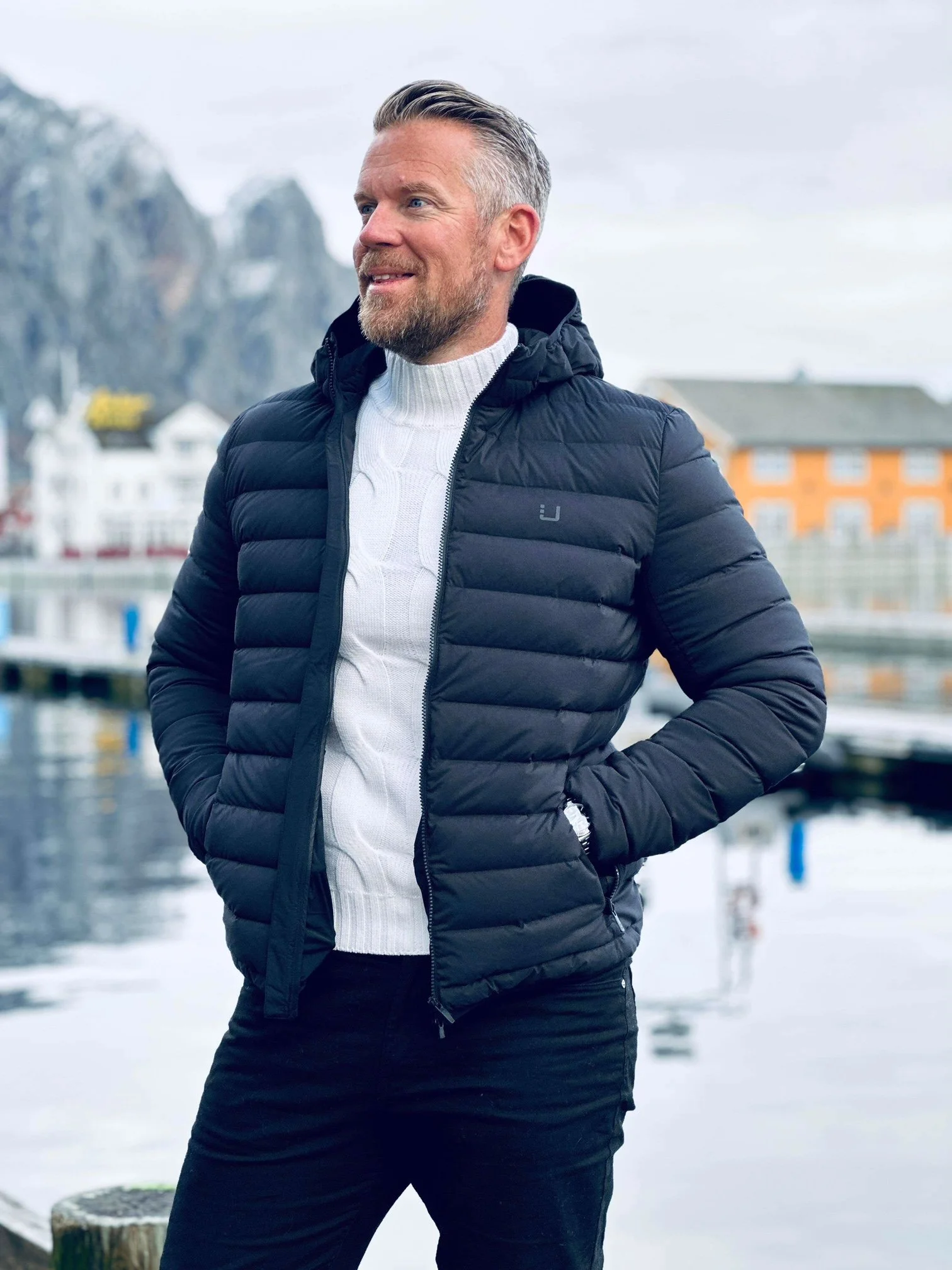 En mann står ved vannet på en sketchy, gråværsdag, iført en svart jakke og en hvit genser.