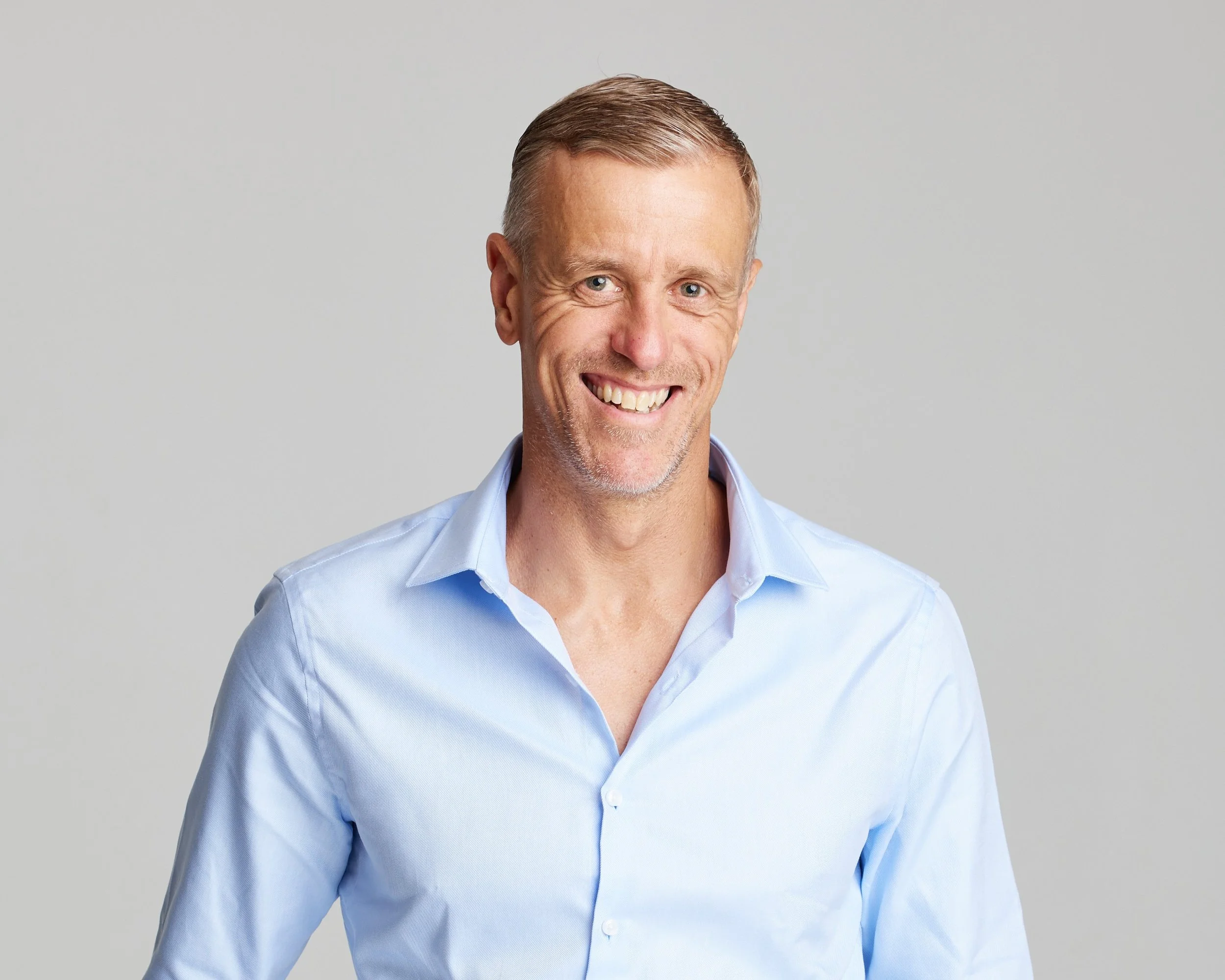 A smiling man with short light brown hair wearing a light blue button-down shirt against a plain gray background.