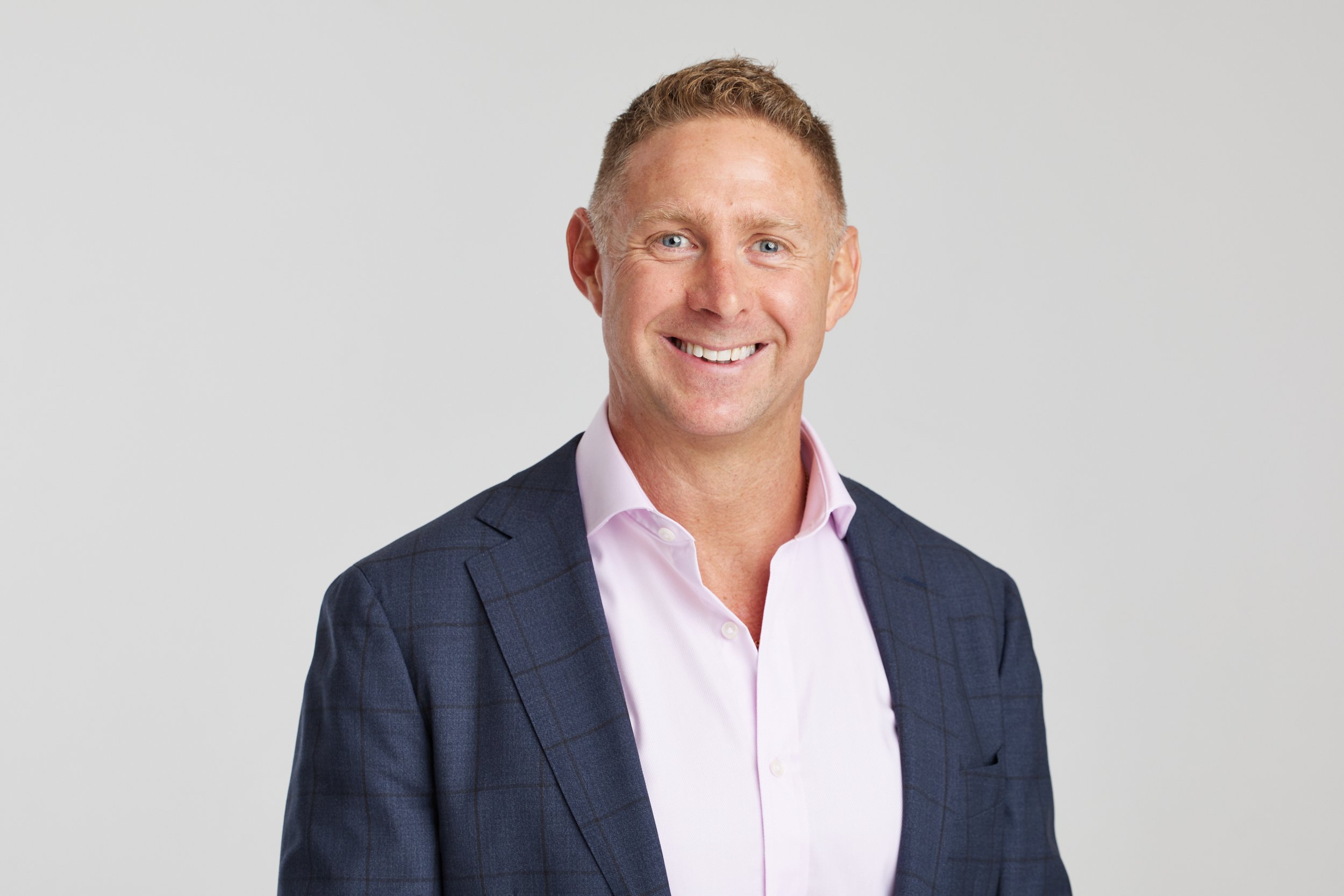 Professional man wearing a dark suit jacket and pink shirt, smiling against a plain gray background.