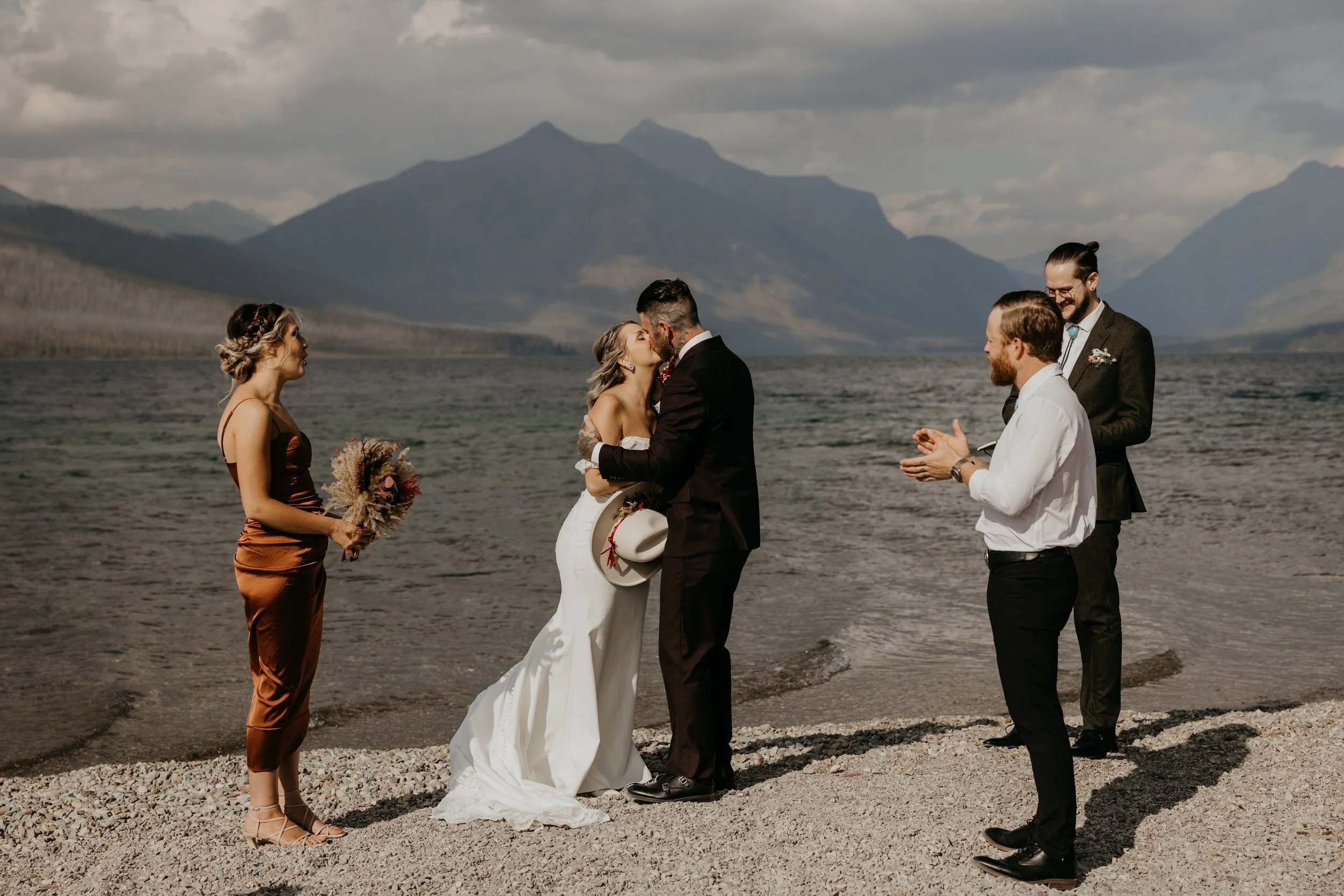 Bride and groom kissing during their elopement ceremony on the shores of Lake McDonald, witnessed by three guests, with mountains in the background, in West Glacier, Glacier National Park.