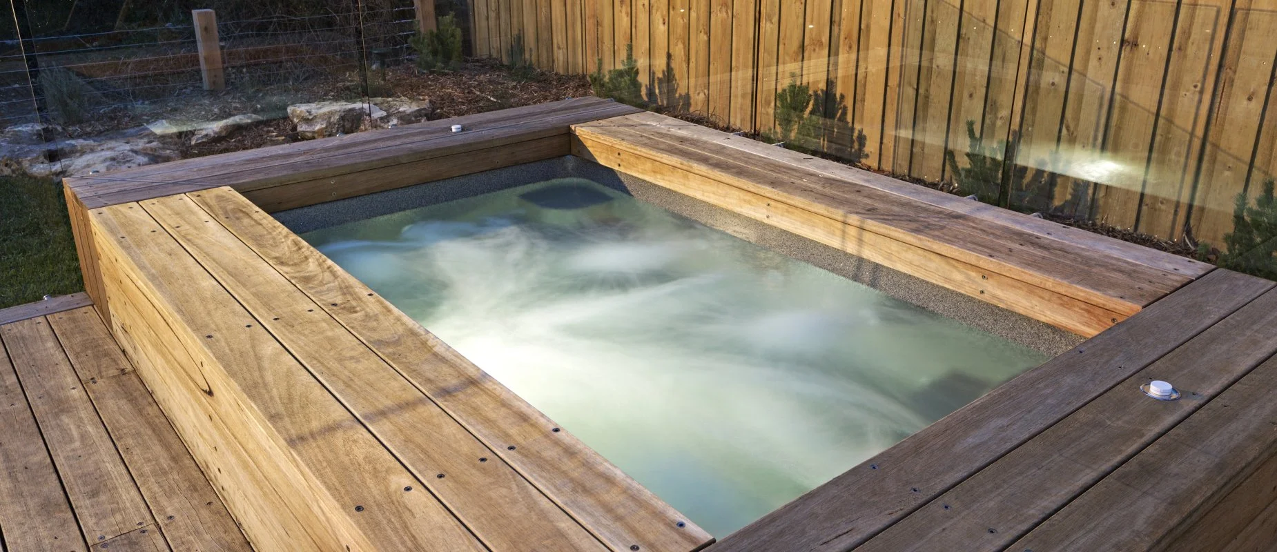 A modern outdoor swimming pool with stepping stones, surrounded by a landscaped area with grass and palm trees. The pool is adjacent to a fenced wall decorated with metal leaf sculptures and colorful lizard artwork.
