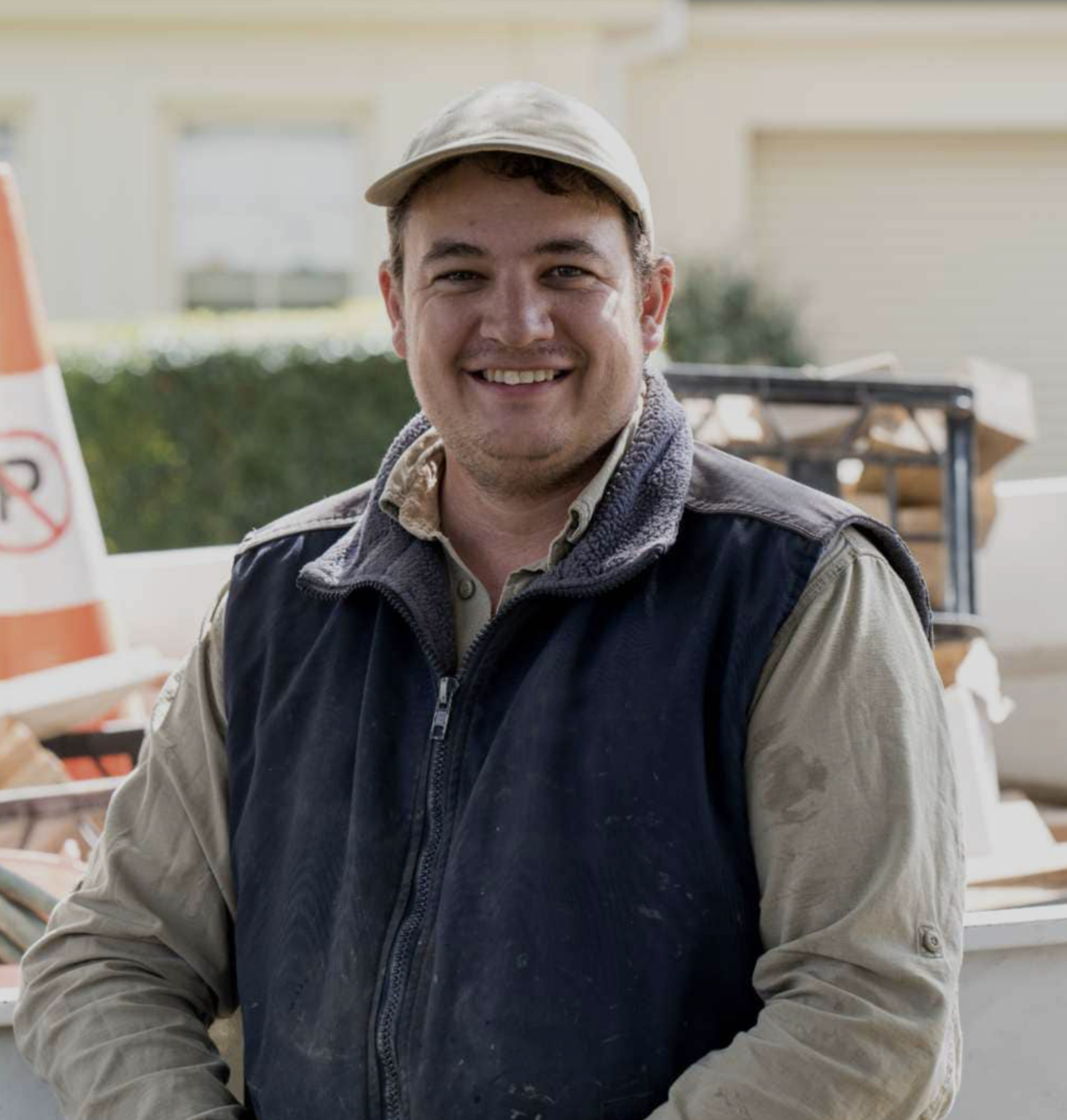 A smiling person wearing a cap and and vest stands outdoors with construction materials in the background.