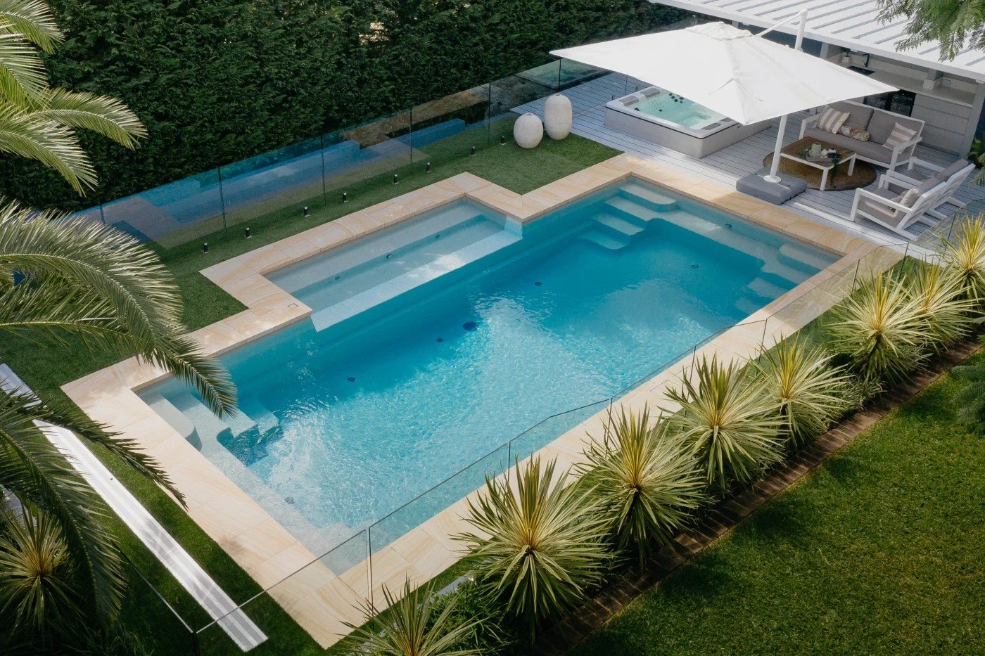Aerial view of a backyard with a rectangular swimming pool surrounded by a glass fence. There is a jacuzzi under a canopy and outdoor seating area with couches and an umbrella. The area is landscaped with palm trees and grass.