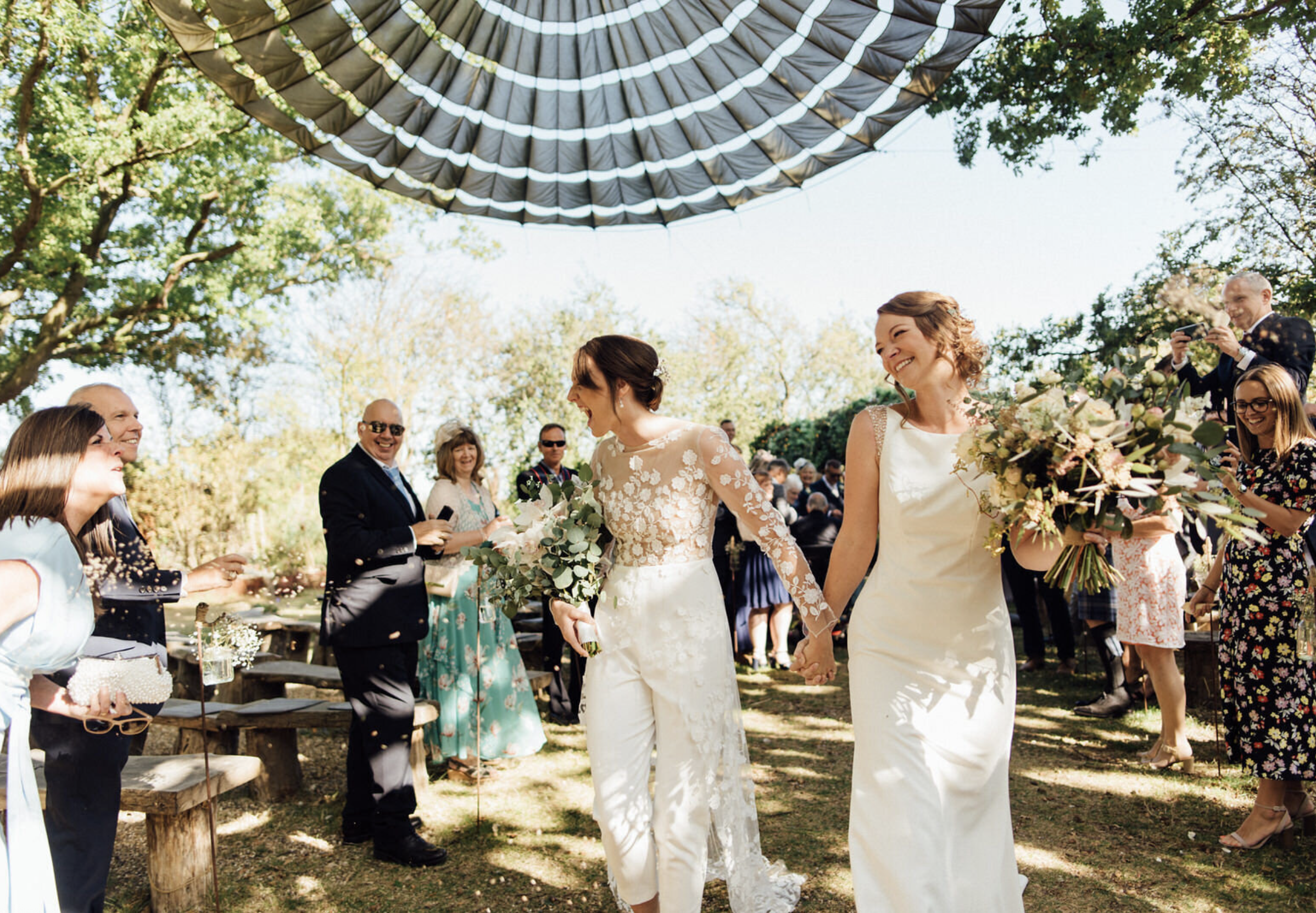 Two women in wedding dresses holding hands and smiling at each other as they walk down the outdoor wedding aisle, surrounded by smiling guests and trees under a large canopy.