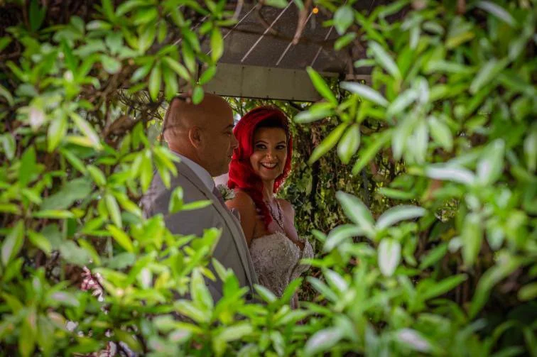 A man and a woman are standing together in a lush garden, partially hidden by green foliage. The woman has red hair and is smiling at the camera while wearing a wedding gown, and the man is dressed in a light gray suit.