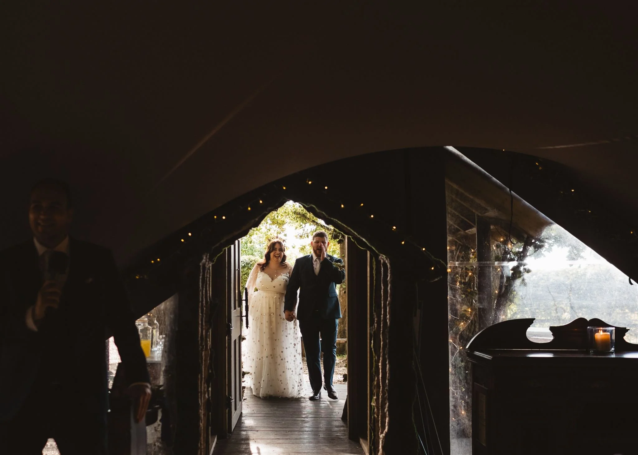 A couple walking into our tent spACE, bride in a white dress and groom in a dark suit, holding hands and smiling.