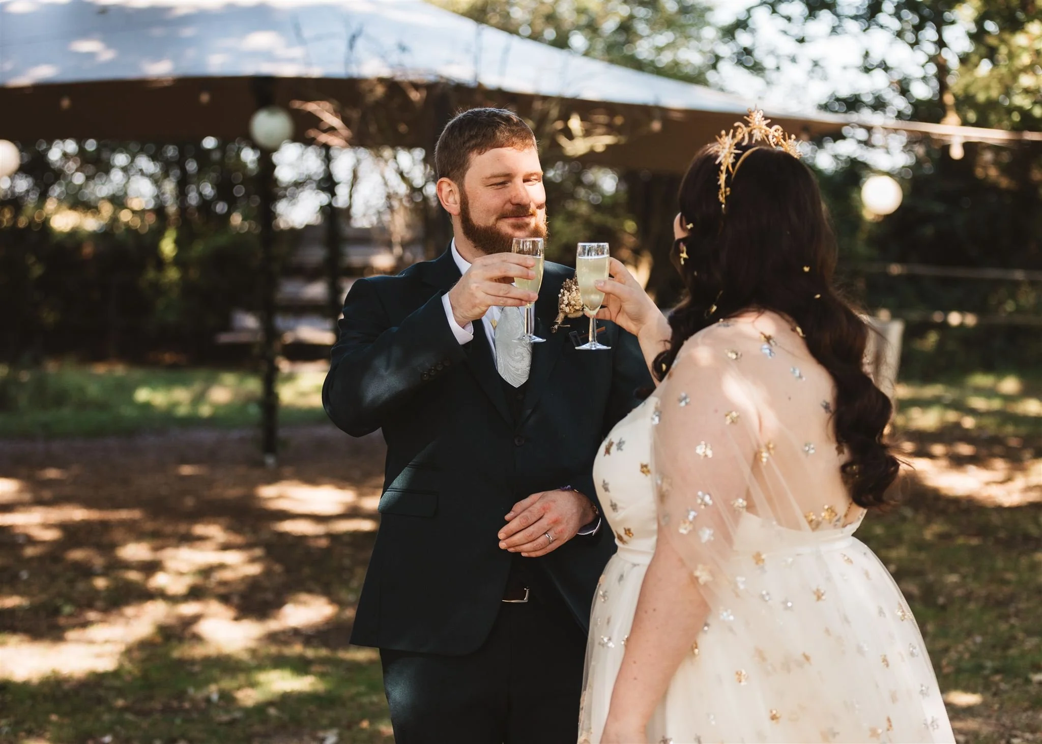 A couple dressed in wedding attire raising glasses of champagne in a toast outdoors with trees and sunlight in the background.