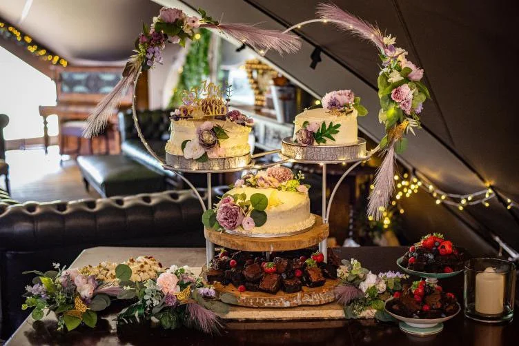 Three white cakes decorated with purple and green flowers and greenery, set on a wooden stand with a decorative metal arch, in a cozy decorated indoor space with fairy lights.