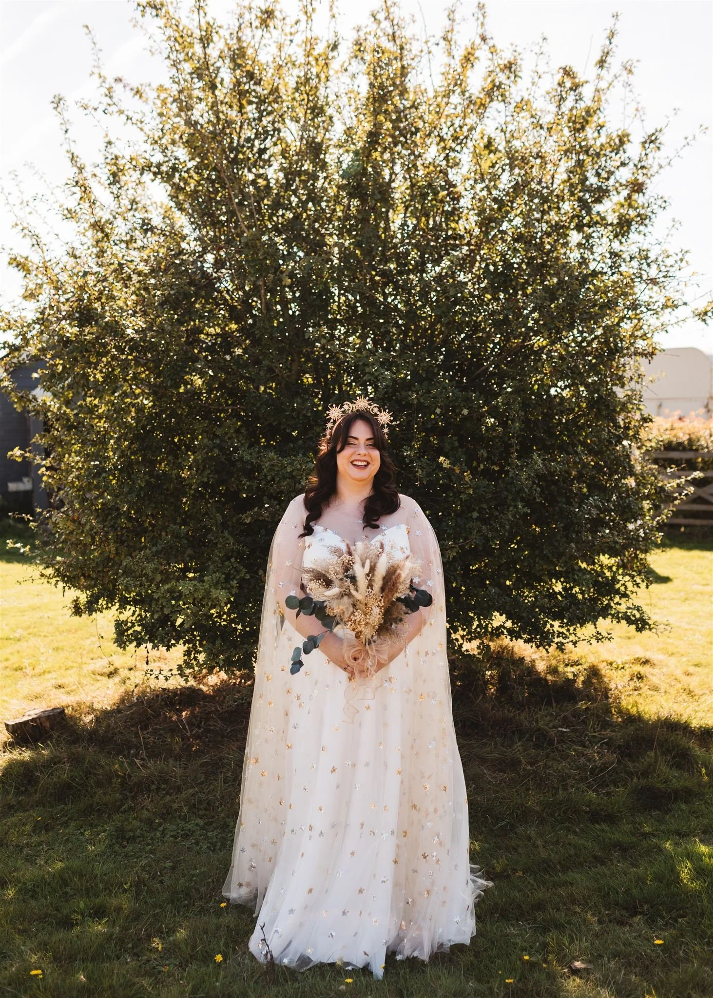 A woman in a white wedding dress holding a bouquet of dried flowers and greenery, standing outdoors in front of a large bush on a sunny day.