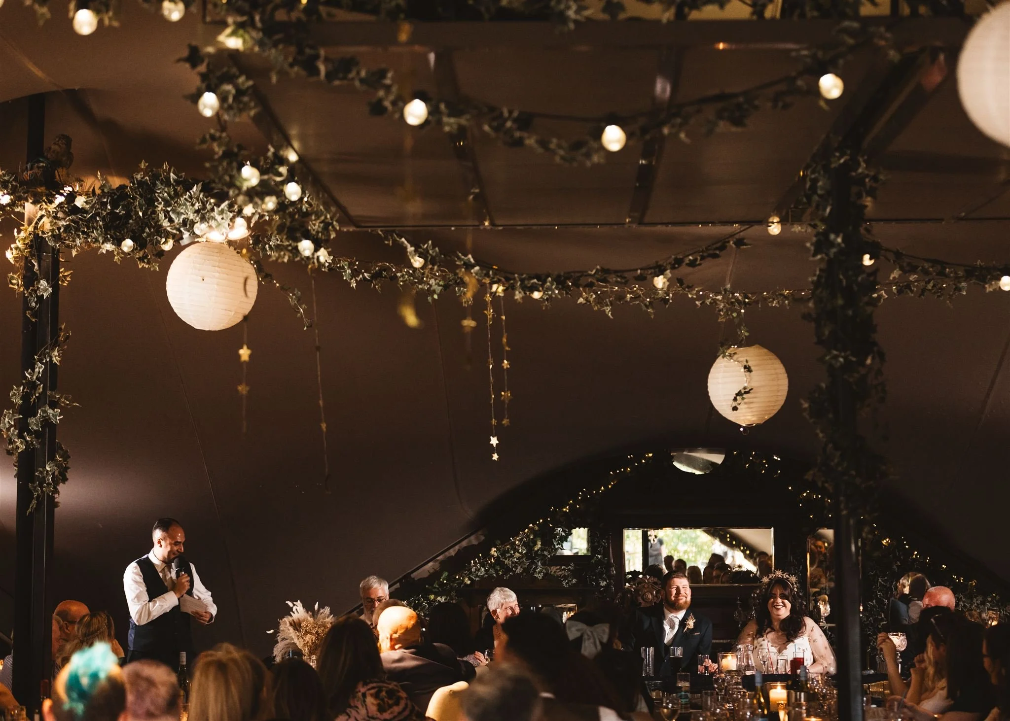 Indoor wedding reception with guests seated at long tables, decorated with candles, flowers, and greenery. A man is giving a speech, standing with a microphone. The ceiling is adorned with hanging paper lanterns, string lights, and greenery garlands.